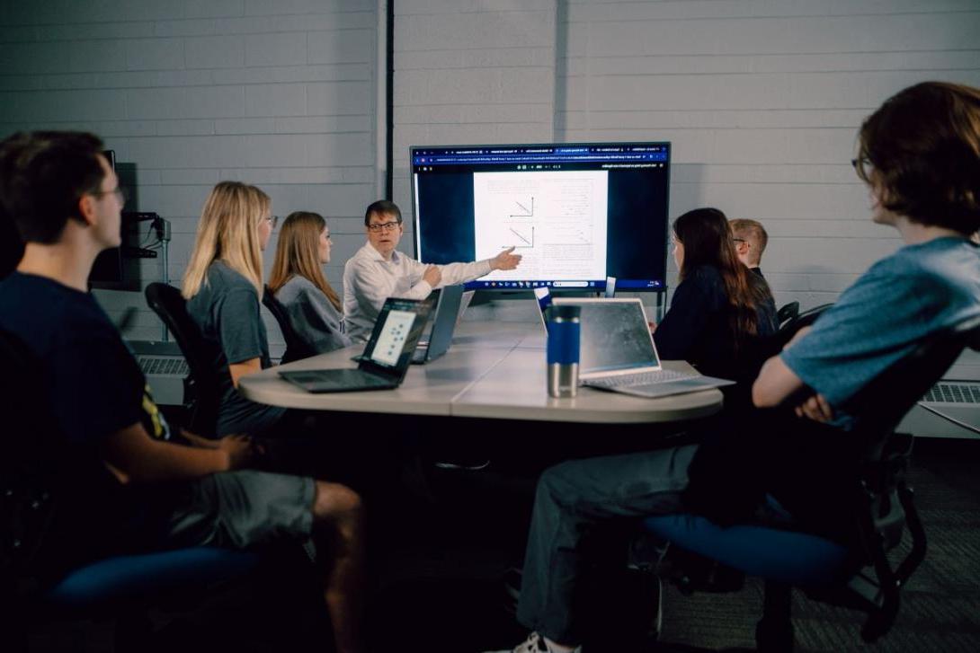 Kettering students watch a professor's computer science presentation. They are seated around a table and have laptops. 